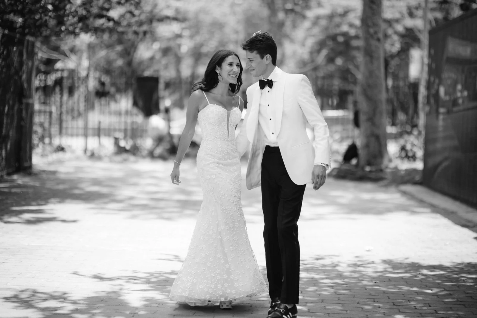 A newlywed couple walking together outdoors, the bride in a white lace wedding gown and the groom in a white tuxedo jacket with black pants and bow tie, smiling and looking at each other.