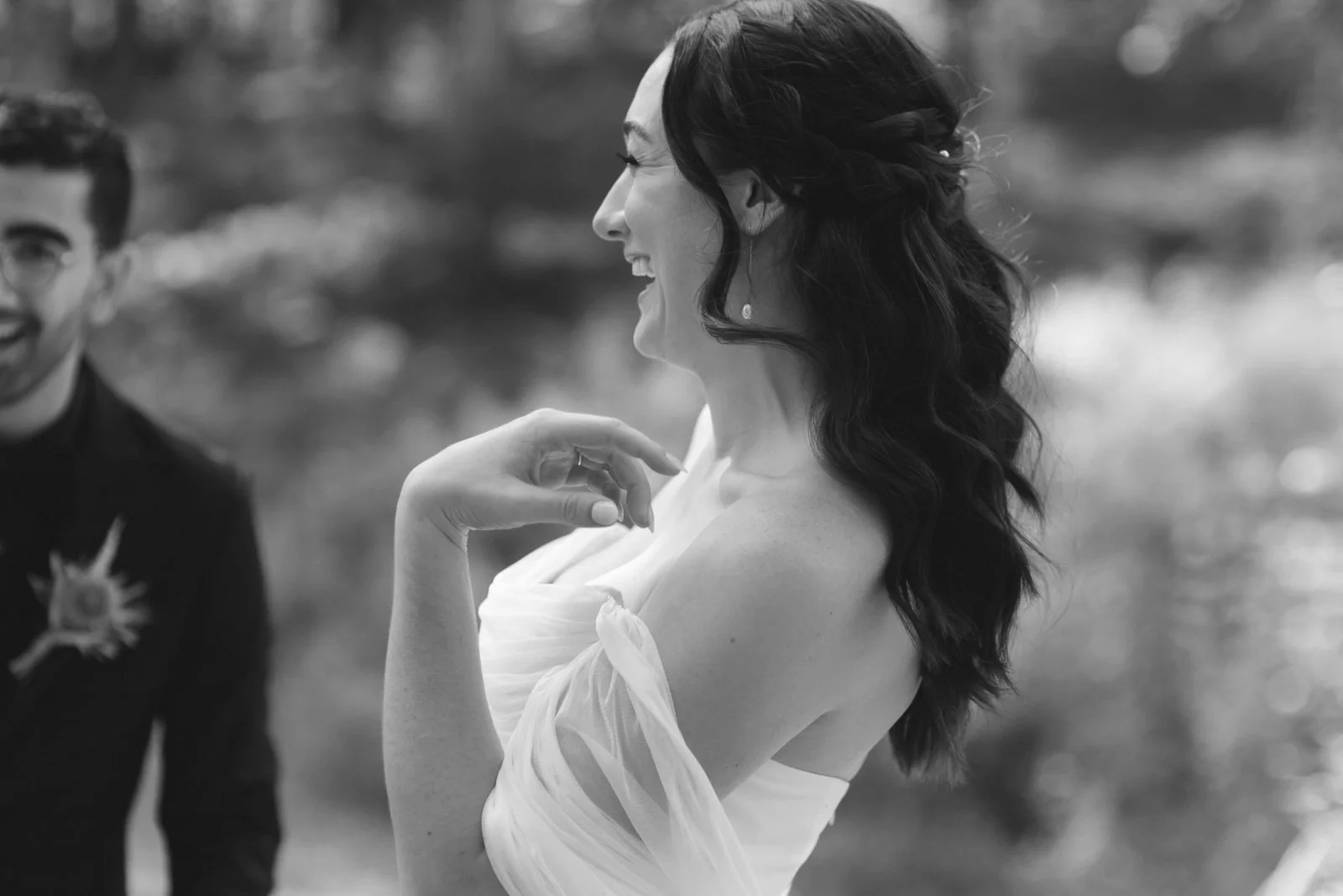 A woman with dark, wavy hair in an elegant dress smiling, with a man in the background, outdoors.