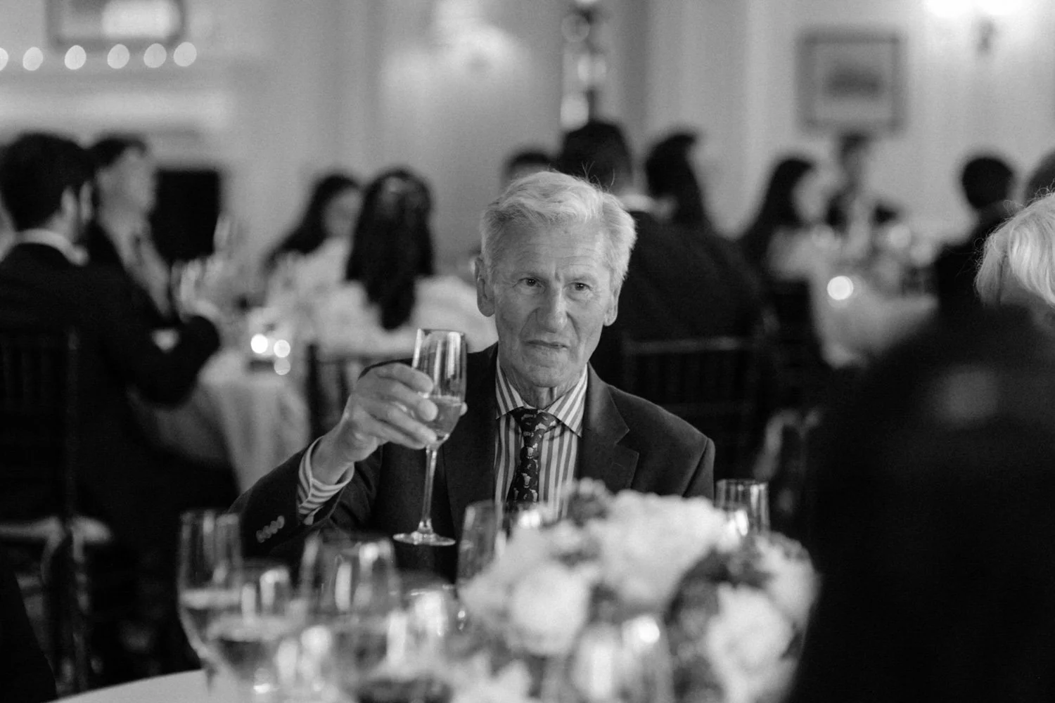 An elderly man in a suit and tie raising a glass at a formal dinner event, with other guests seated at tables in the background.