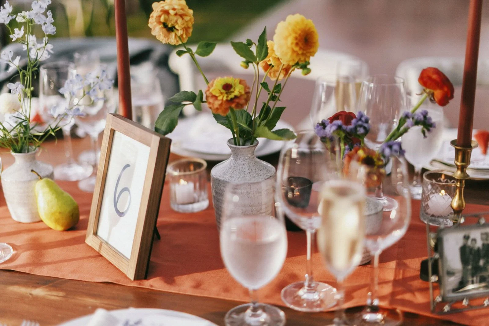 A table decorated for an event with floral arrangements in vases, candles, a framed table number, and drinking glasses. There is a pear and a green apple next to the table number.