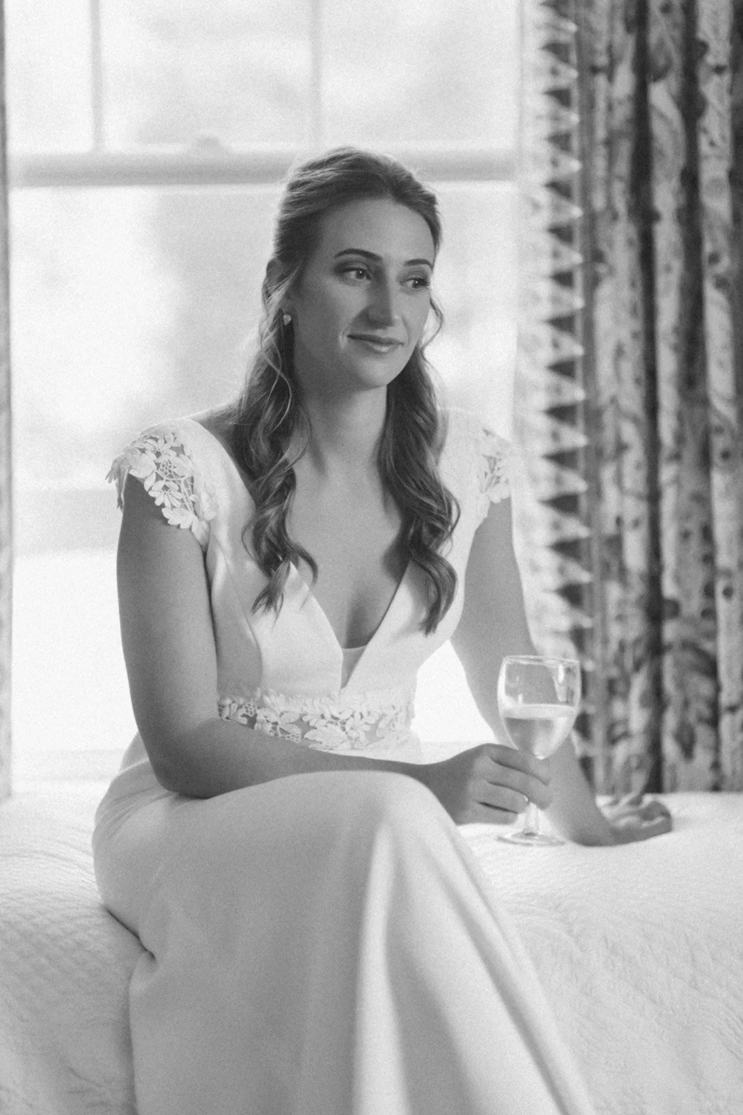 A woman in a white dress with lace details on the shoulders and waist, sitting on a bed, holding a glass of white wine, with patterned curtains in the background.