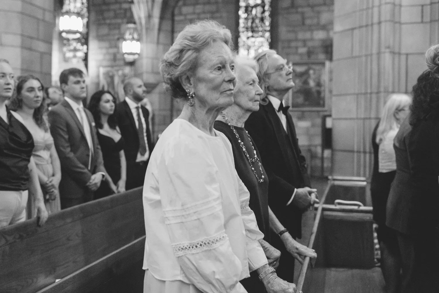A group of people attending a church service, with elderly women standing at the front and younger individuals behind them, inside a church with brick walls and stained glass windows.