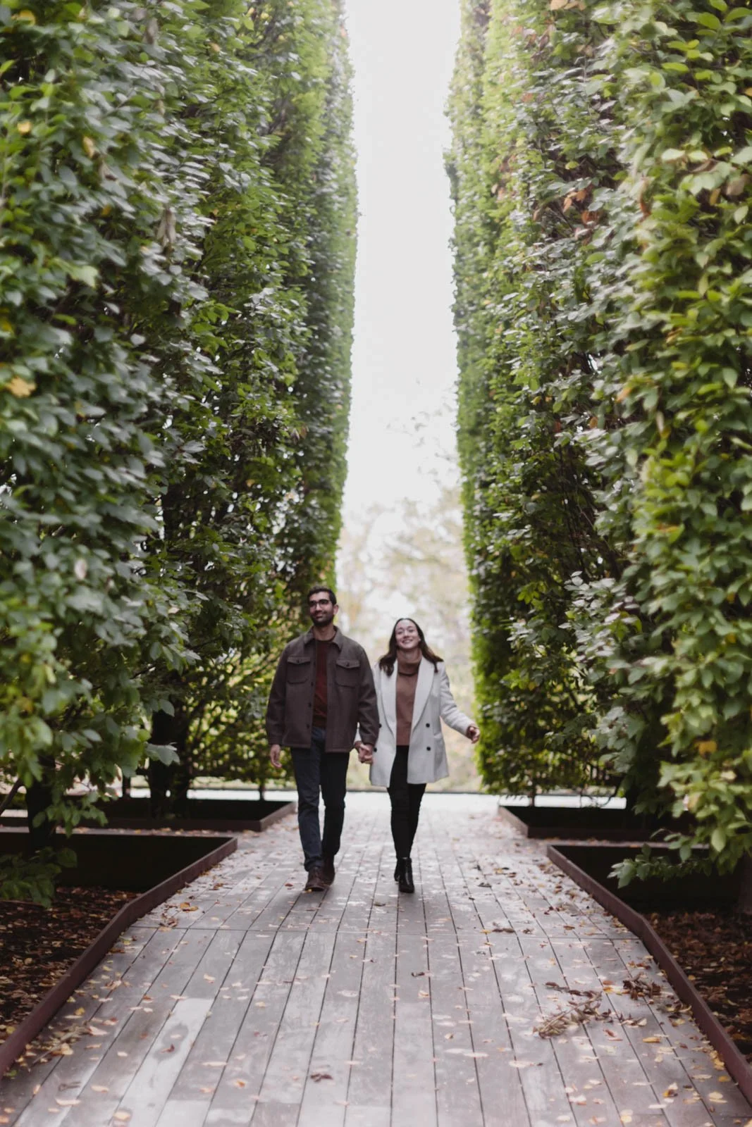 A couple walking hand in hand on a wooden pathway surrounded by tall, lush green hedges in an outdoor garden at DIA Beacon.