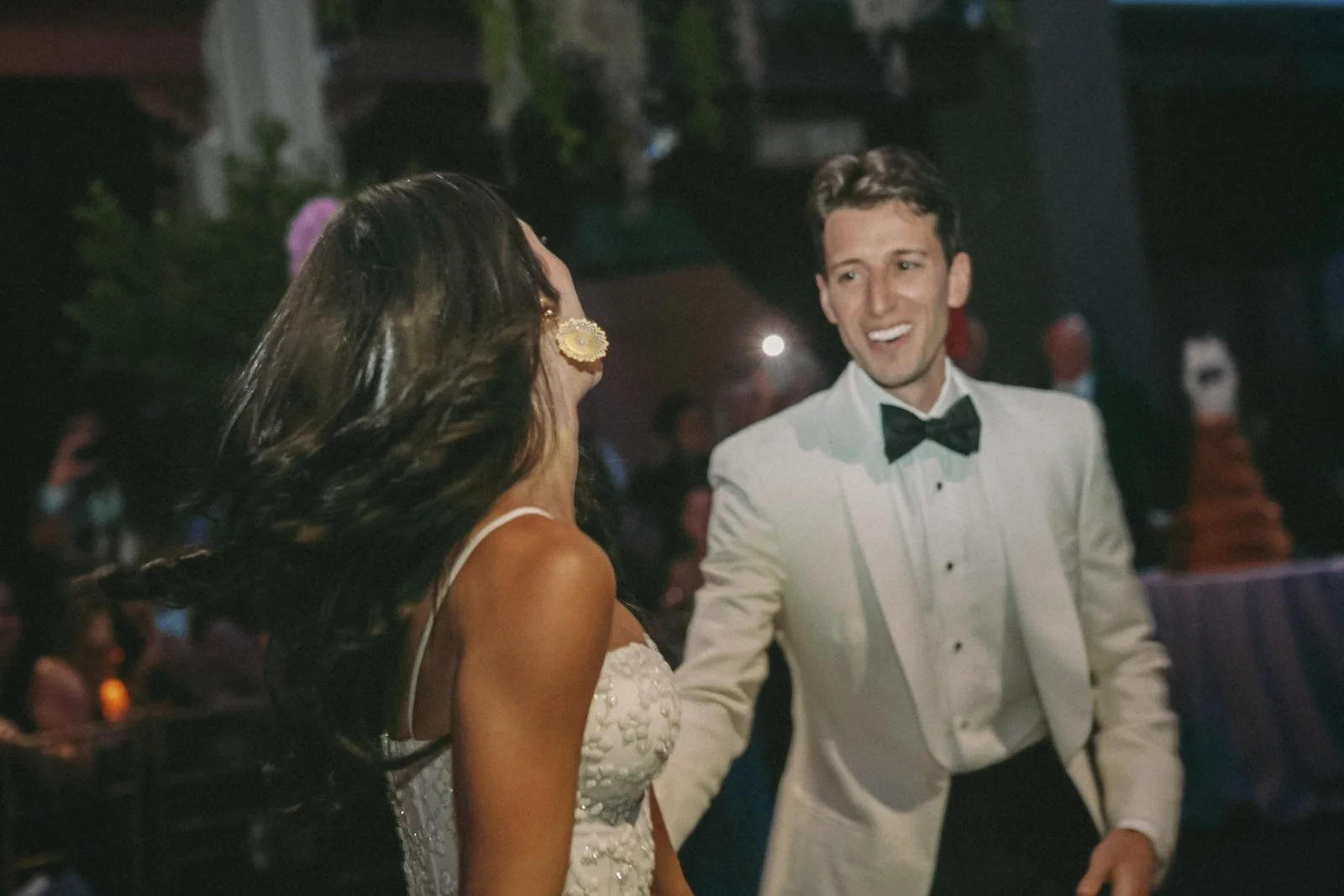 A young man in a tuxedo dancing with a woman in a white dress at a celebration or wedding reception, with others in the background.