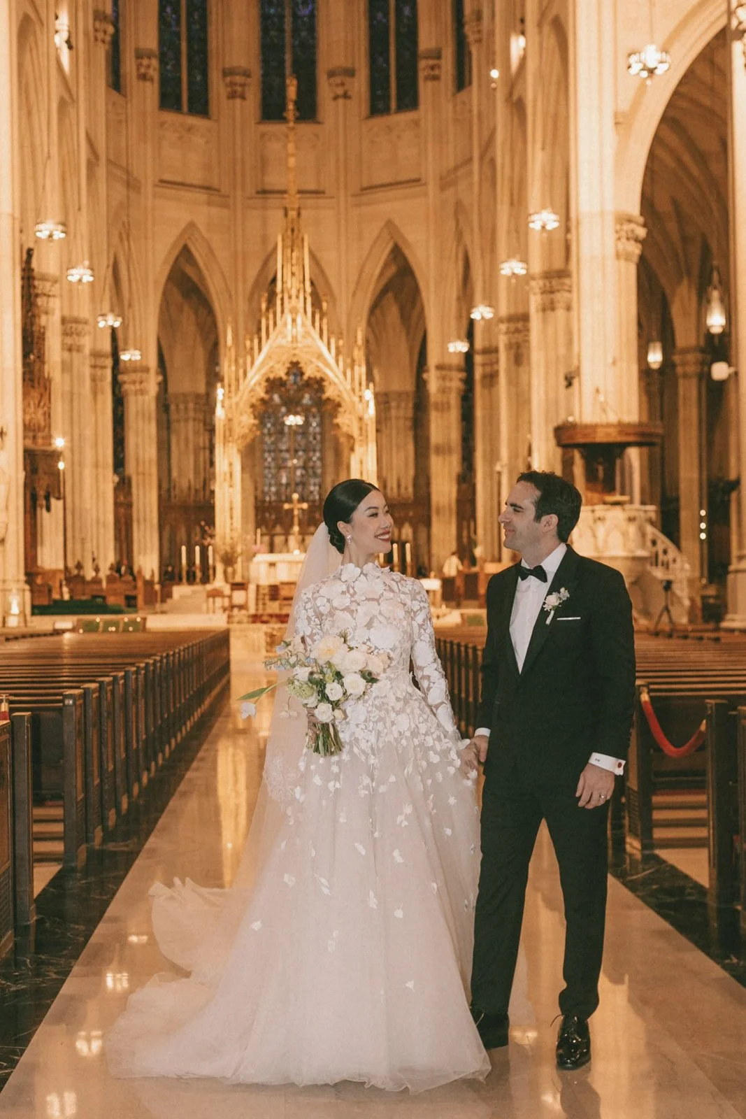Bride and groom walking hand in hand inside a grand, ornate church, smiling at each other. The bride wears a white lace wedding gown and holds a bouquet, while the groom is dressed in a black tuxedo with a bow tie. The church has high vaulted ceiling