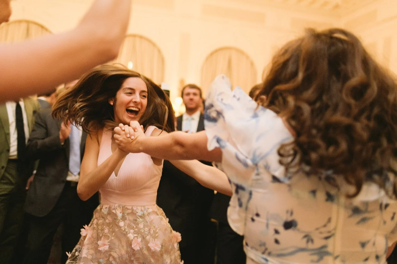 Two women dancing and having fun at a celebration, holding hands, smiling, with people in formal attire in the background.
