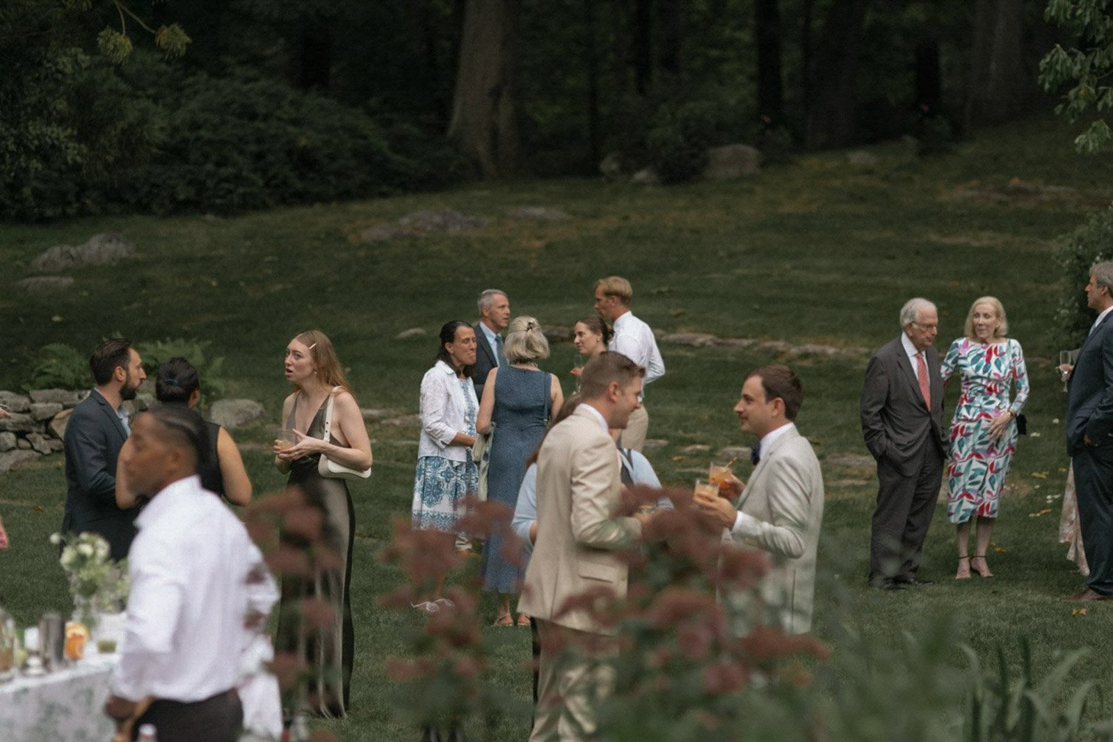 People socializing at an outdoor event in a grassy, wooded area during the evening.