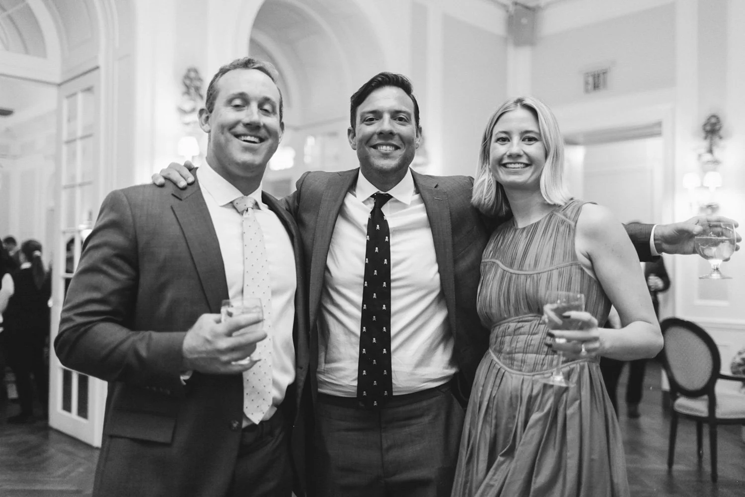 Three people at a social event, smiling, holding drinks, with two men in suits and a woman in a dress, standing in an elegant room.