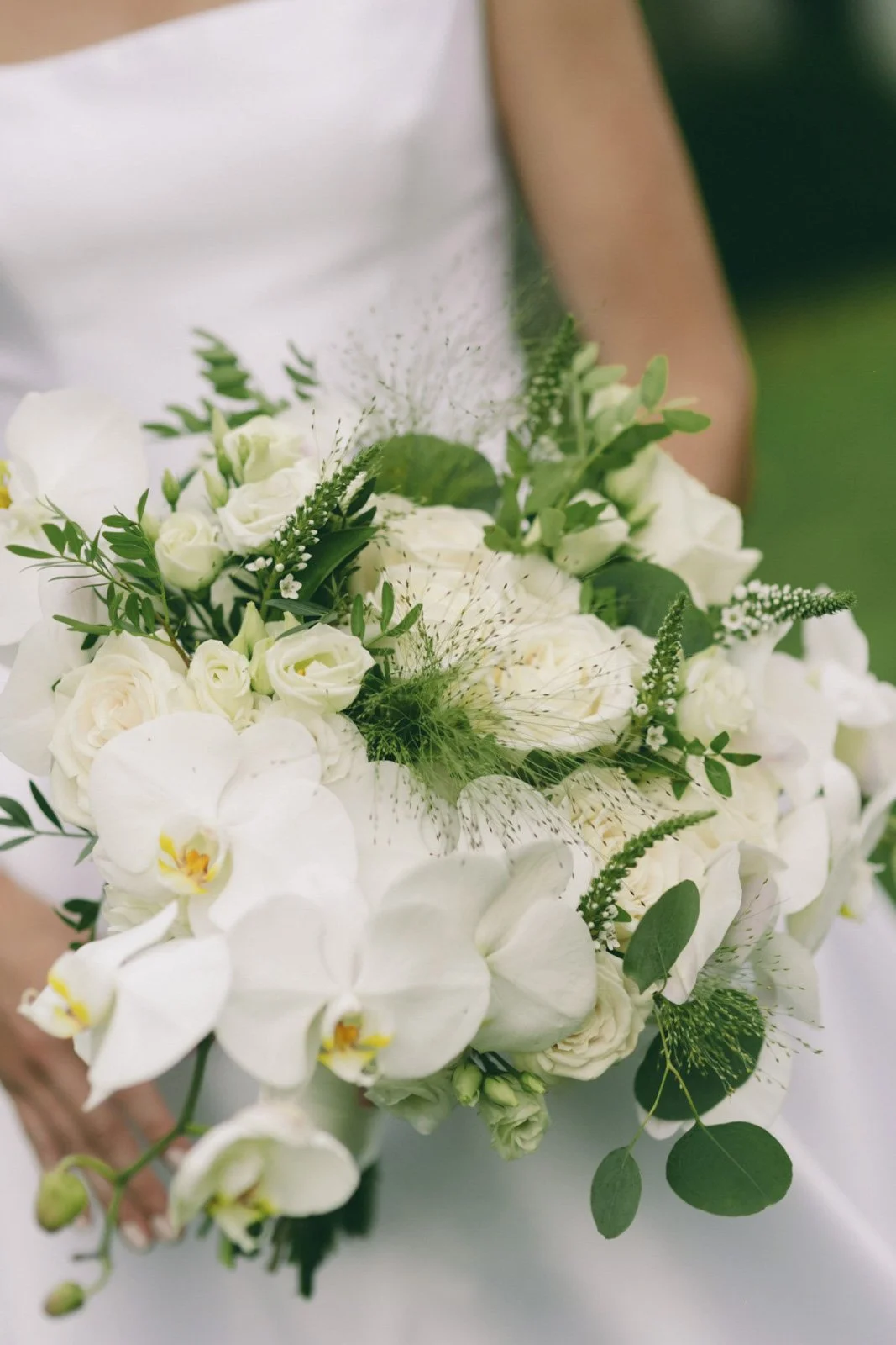 Close-up of a bouquet of white flowers including orchids, roses, and greenery, with a blurred background.