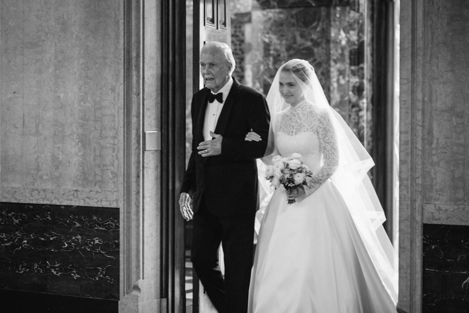 A bride in a wedding dress holding a bouquet being escorted by an older man in a tuxedo at the entrance of a building.