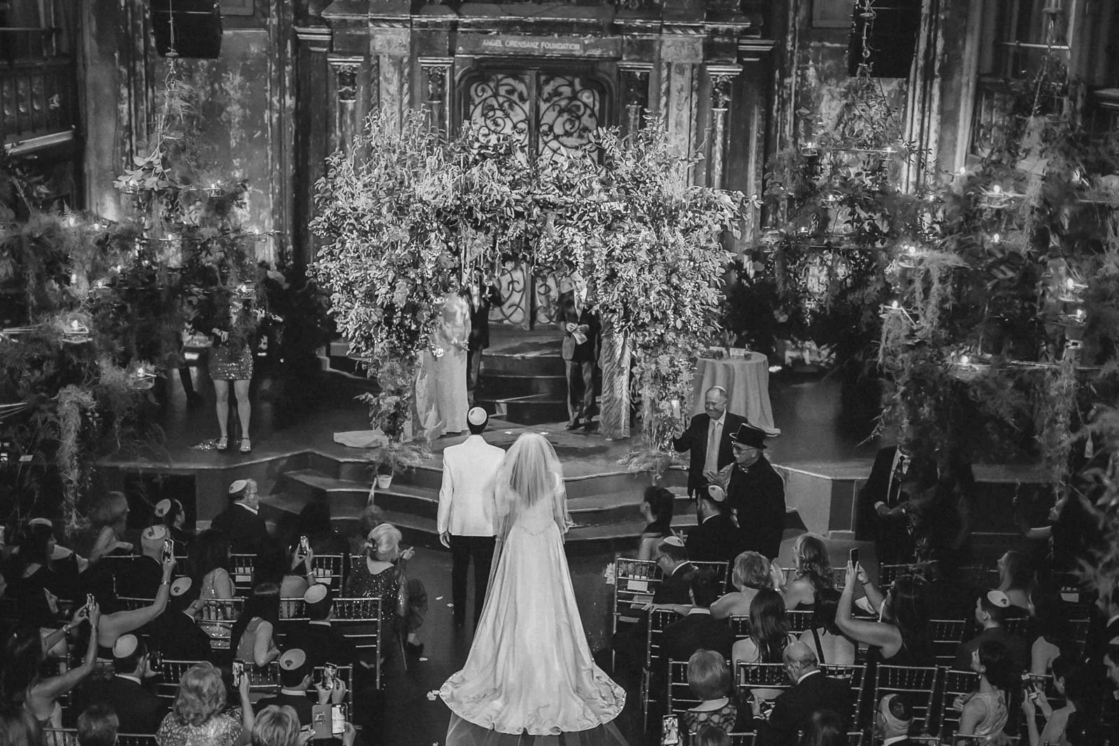 A wedding ceremony taking place in a decorated church with a bride and groom standing at the altar with guests seated around.