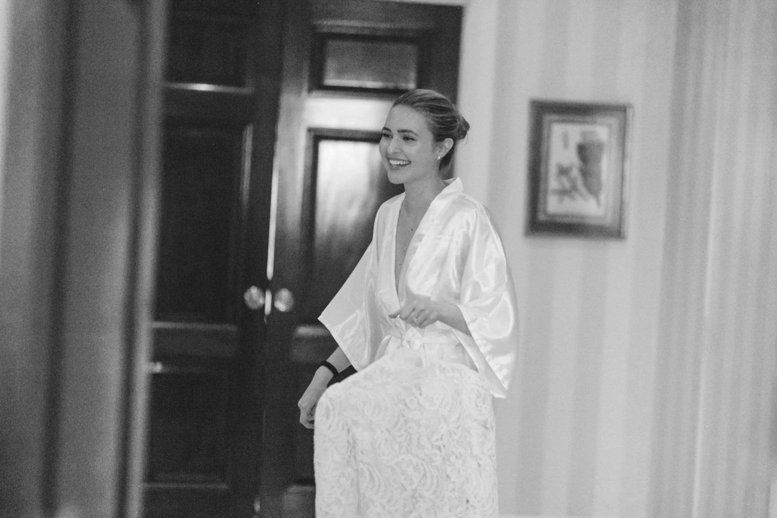 A smiling woman in a satin robe standing in a room with wooden furniture and framed artwork on the wall.