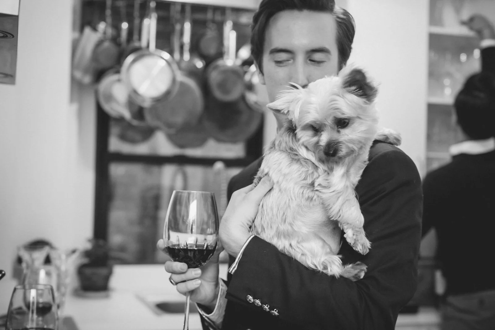 A man in a suit holding a small dog and a glass of red wine in a restaurant.
