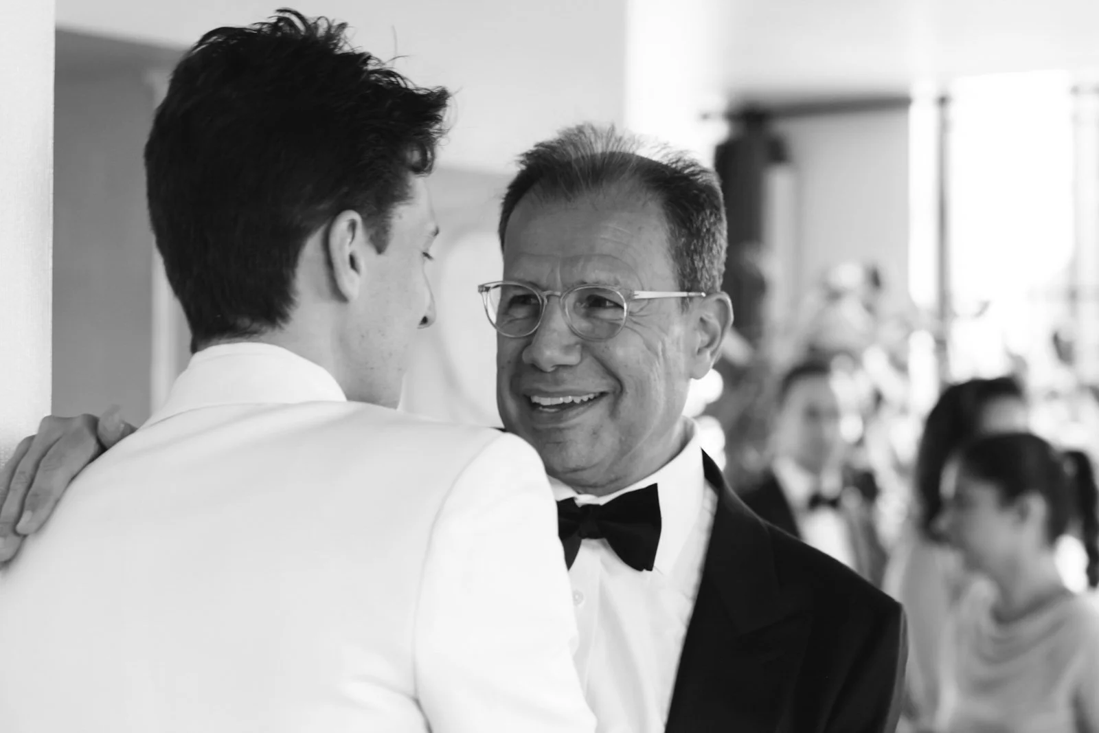 Two men, one younger and one older, in formal attire smiling and embracing at a social event.