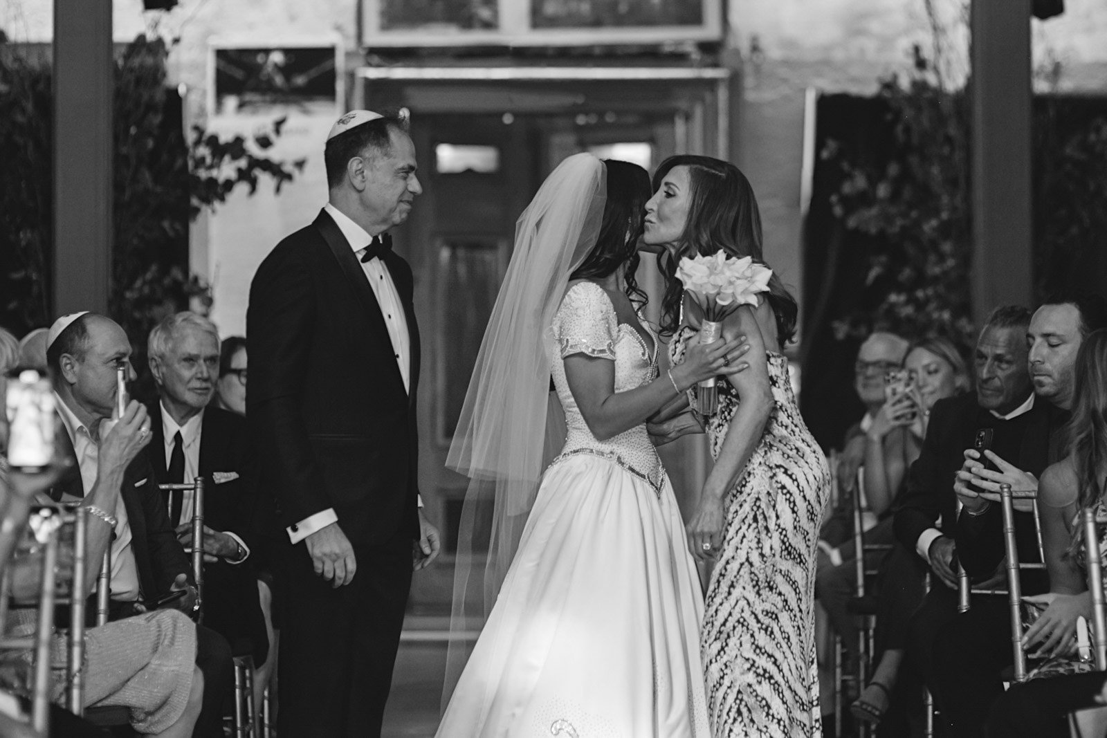 A black and white photo of a wedding ceremony showing a bride in a wedding dress, holding a bouquet of flowers, leaning in to kiss a woman in patterned dress, while a man in a tuxedo and kippah looks on. Guests are seated on either side, some taking 