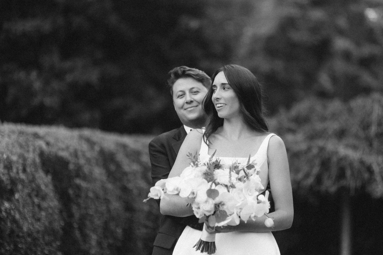 A black and white photograph of a bride holding a bouquet of flowers with a groom standing behind her, outdoors with trees in the background.