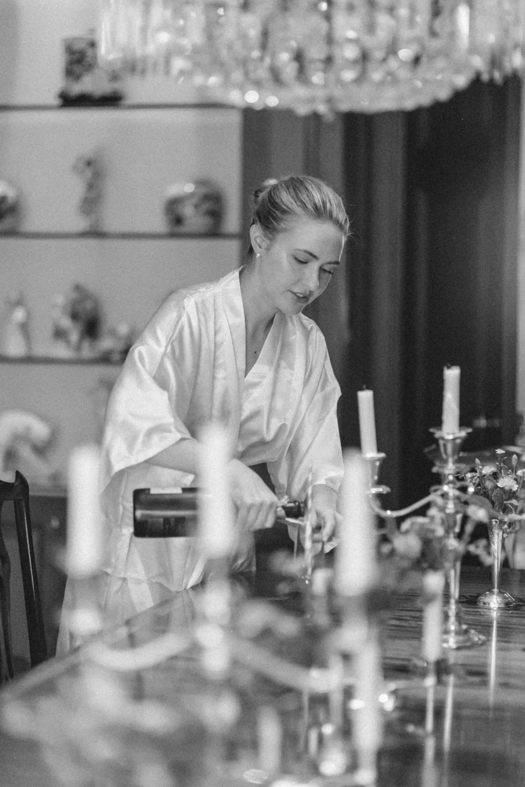 A woman in a satin robe decorating a table with candles and flowers as she lights a candlestick.