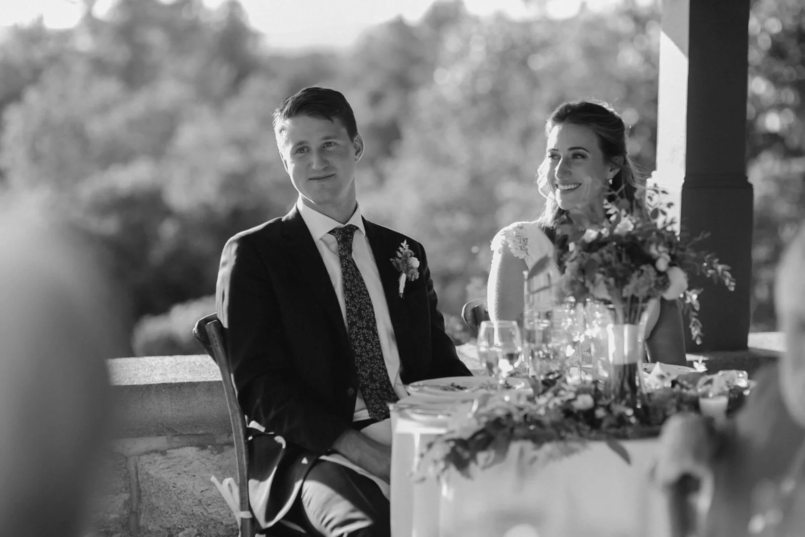 A black and white photo of a wedding reception with a bride and groom sitting at a decorated table outdoors, smiling and looking at the officiant or guest.