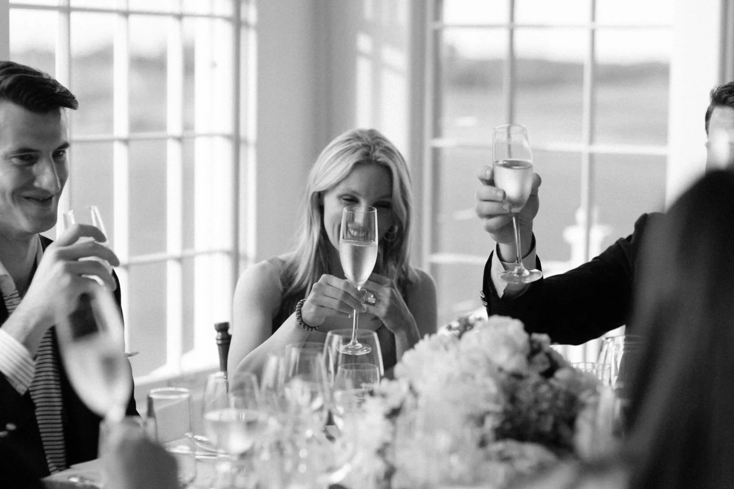 People celebrating with glasses of champagne at a banquet table, in a bright room with large windows.