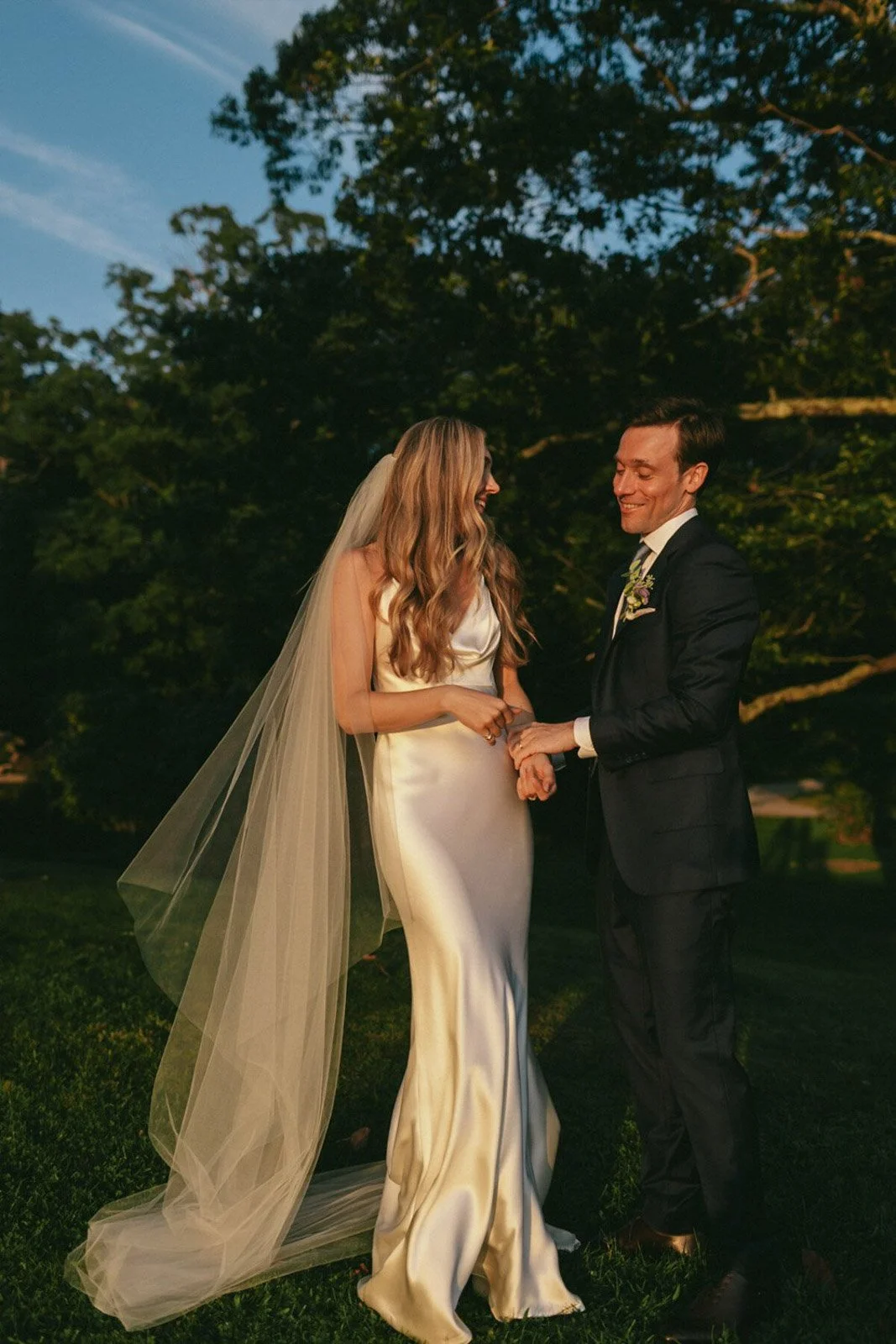A bride and groom holding hands and smiling at each other in an outdoor setting during their wedding at sunset, with trees in the background.