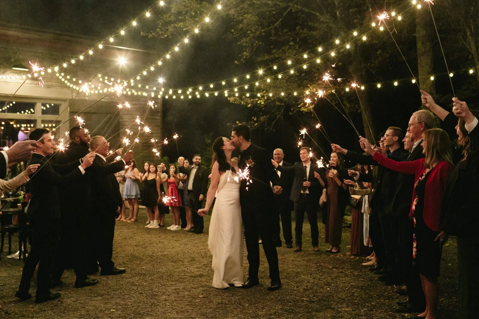 A bride and groom kiss in the center of a nighttime outdoor wedding celebration, surrounded by guests holding sparklers. String lights hang above, creating a festive atmosphere.