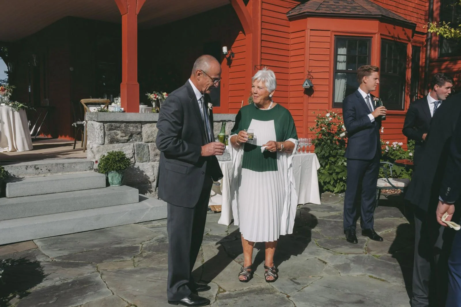 People at an outdoor wedding reception, talking and holding drinks, with a red house and decorated tables in the background.