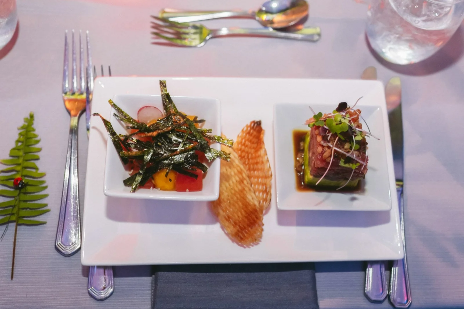 A white rectangular plate with two small square dishes containing sashimi and salad, a crispy rice chip, placed on a gray tablecloth with silverware, a glass of water, and a fern leaf with a ladybug to the left.