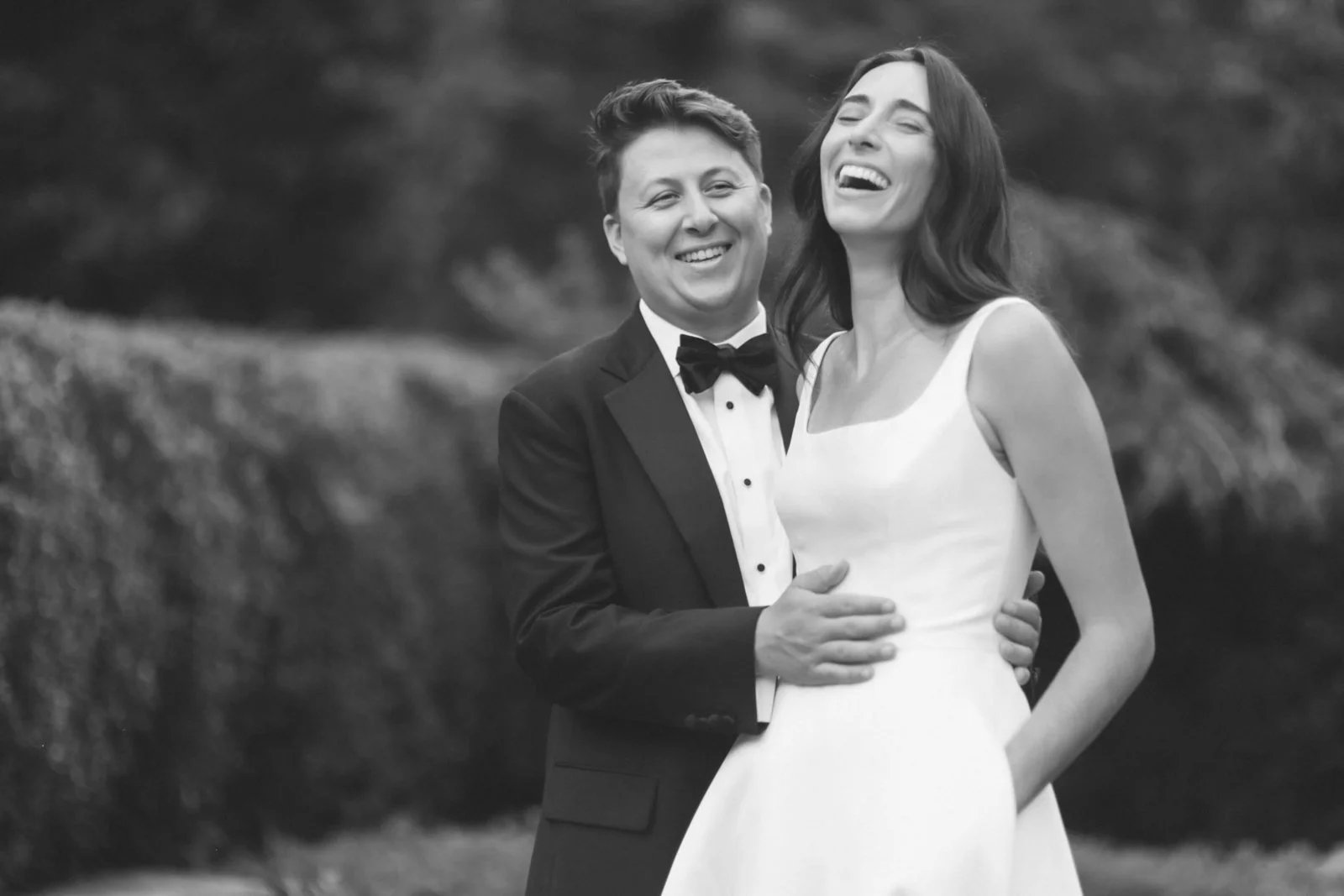 A black-and-white photo of a happy couple in formal wedding attire, with the man in a tuxedo and bow tie, and the woman in a white wedding dress. They are smiling and embracing outdoors.