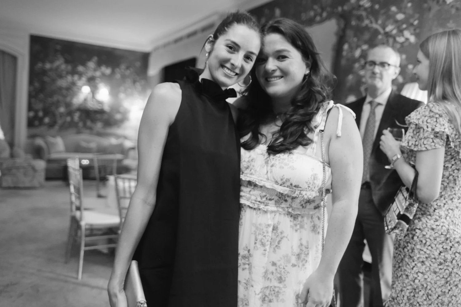 Two women smiling and posing for a photo at a social gathering, with other guests in the background.