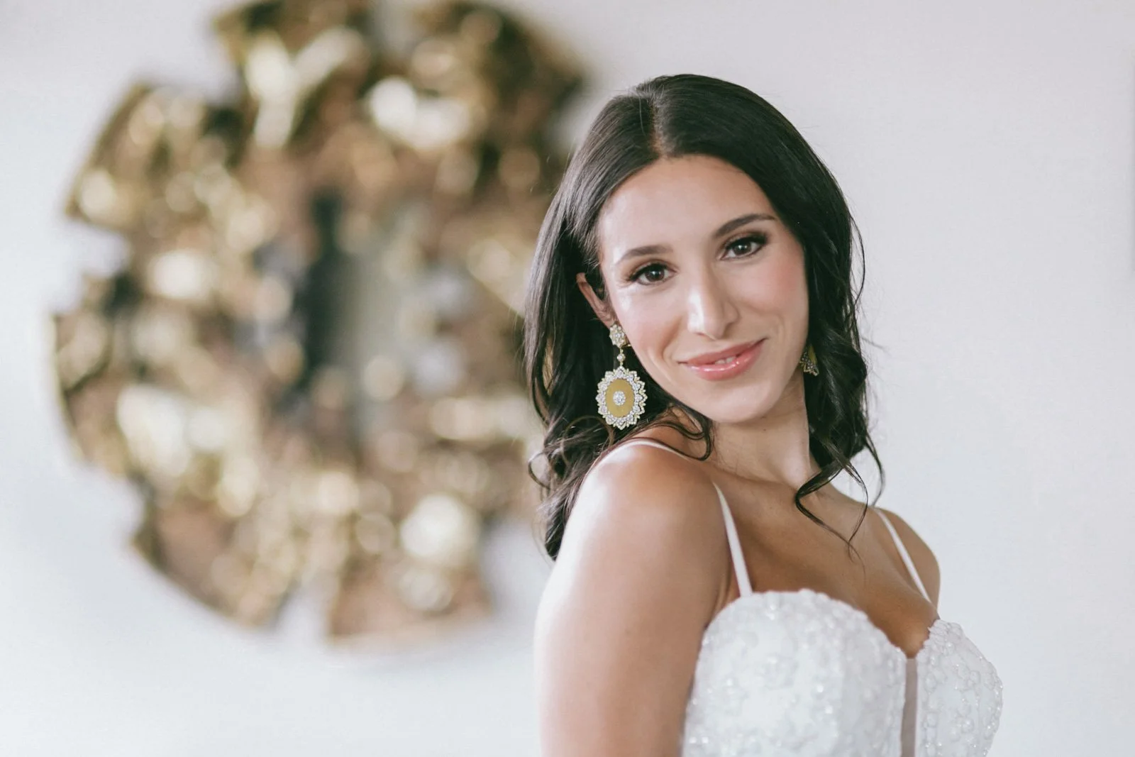 A young woman with dark hair smiling, wearing a white dress with thin straps and large, ornate earrings, against a white background with a decorative gold mirror or artwork behind her.