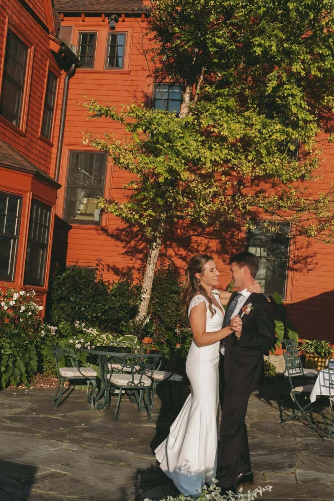 A bride and groom sharing a romantic dance in a garden with lush greenery and flowers, adjacent to a red wooden building, during an outdoor wedding celebration on a sunny day.
