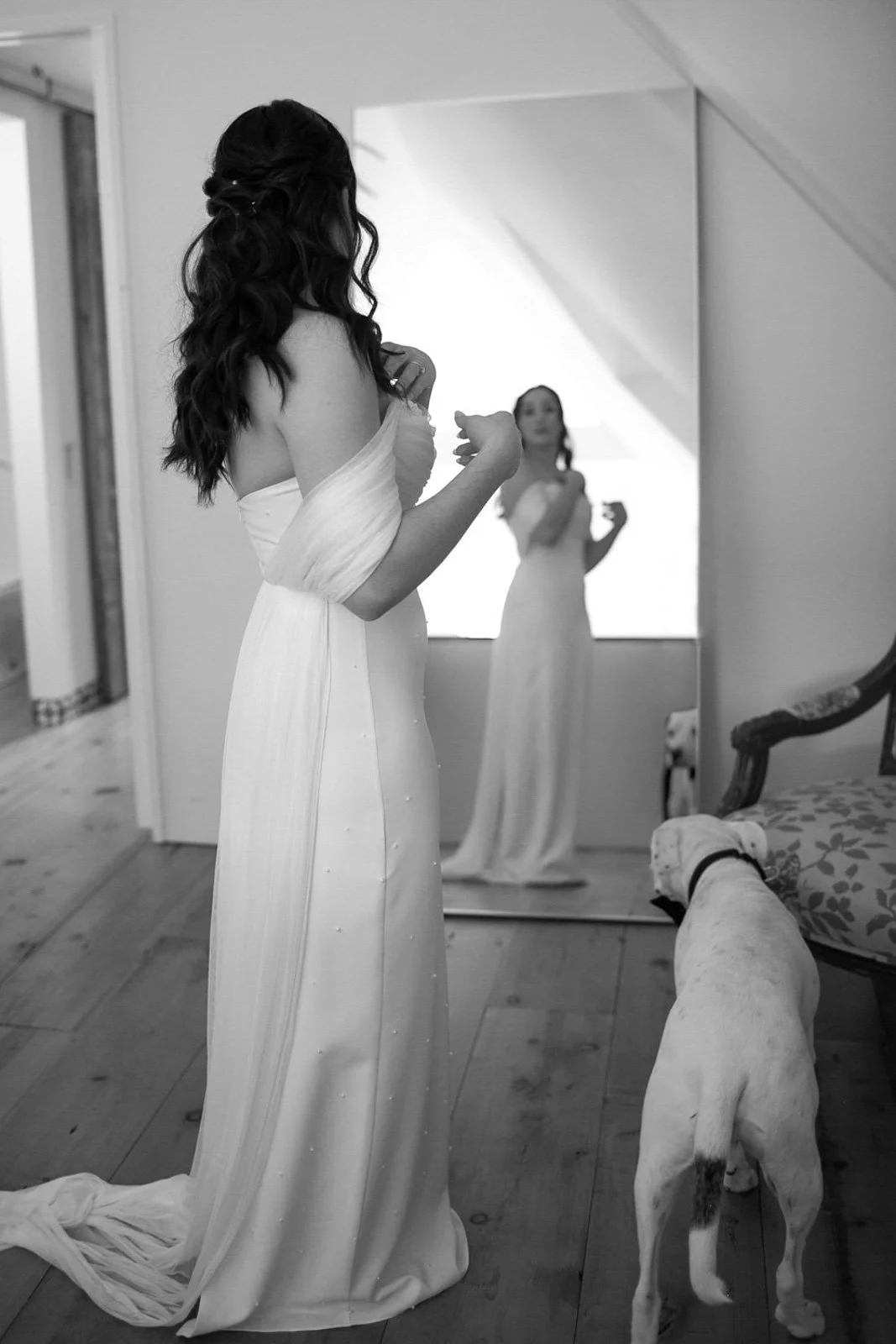 A bride in a wedding dress is looking at herself in a mirror, with a dog nearby in a room with wooden floors and furniture.