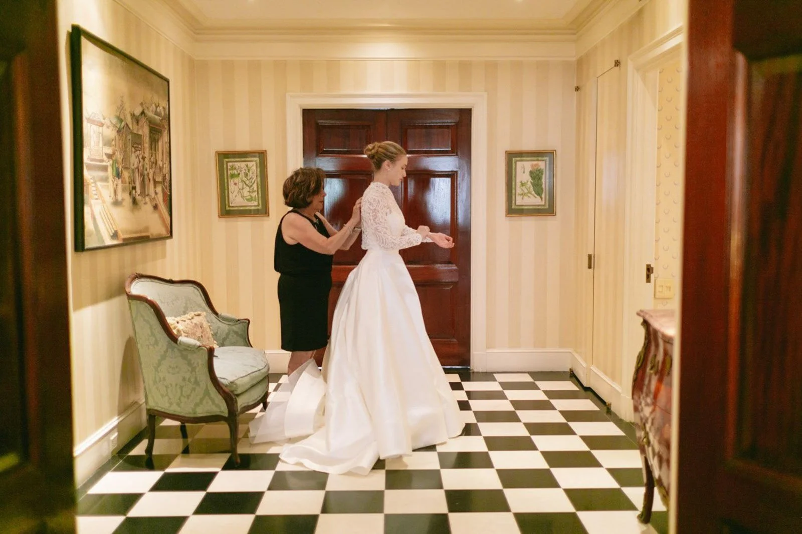 A bride in a white wedding dress standing in a hallway with yellow striped wallpaper, a black and white checkered floor, and a woman helping her with her dress.