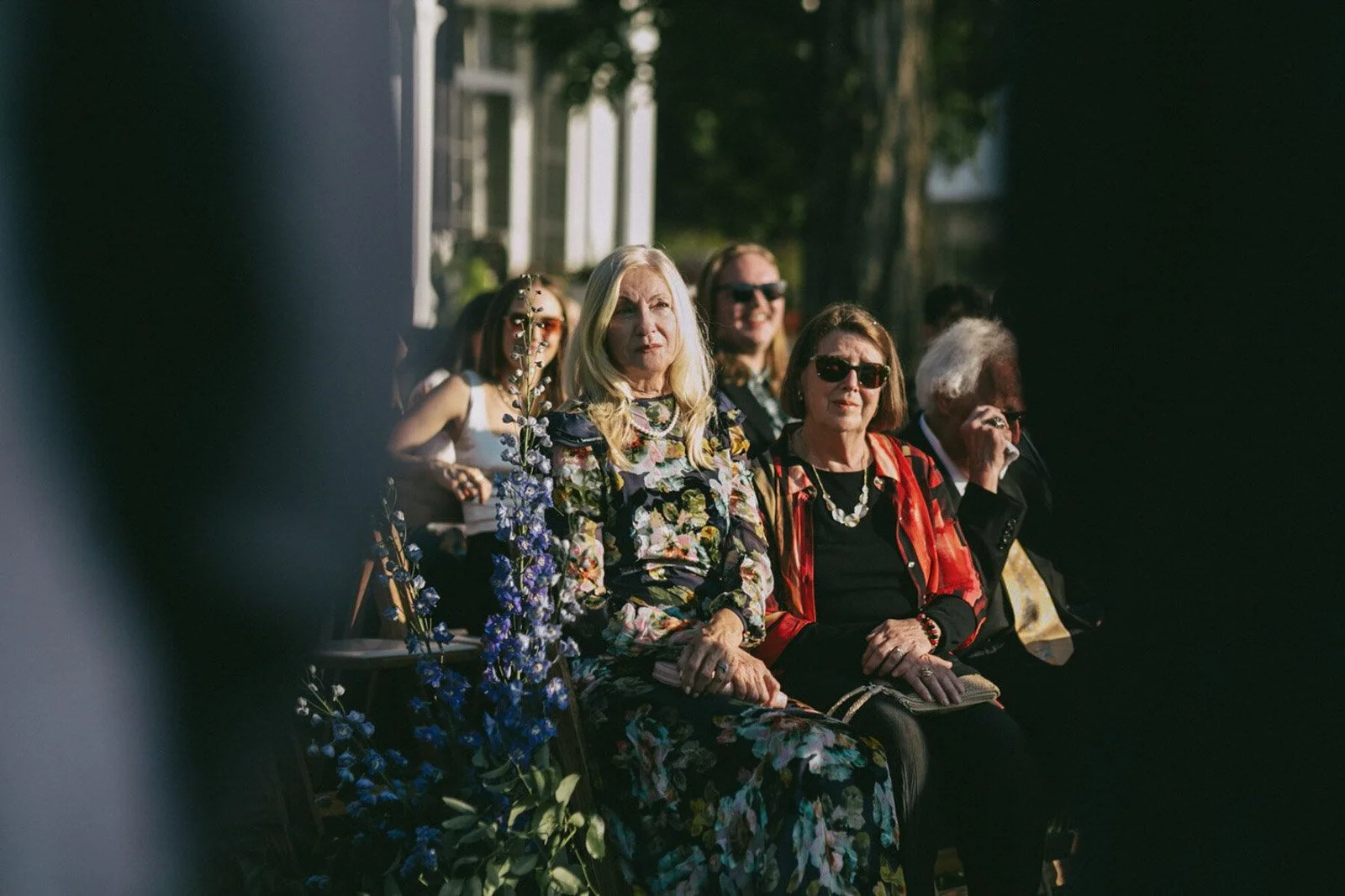 A group of people sitting outdoors, watching an event, with trees and a building in the background. The focus is on a woman with blonde hair and a floral dress, alongside others wearing sunglasses and casual attire.
