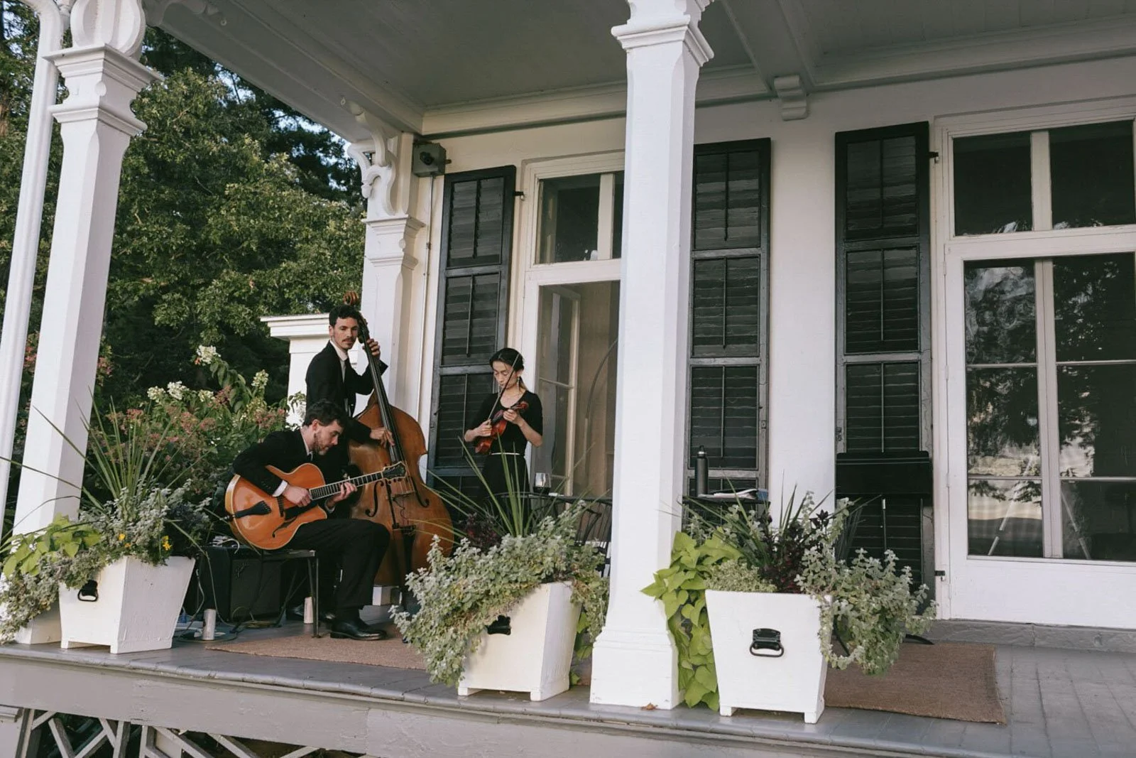 Musicians performing on a porch, with one playing guitar, another on upright bass, and a third playing violin, surrounded by flower arrangements.