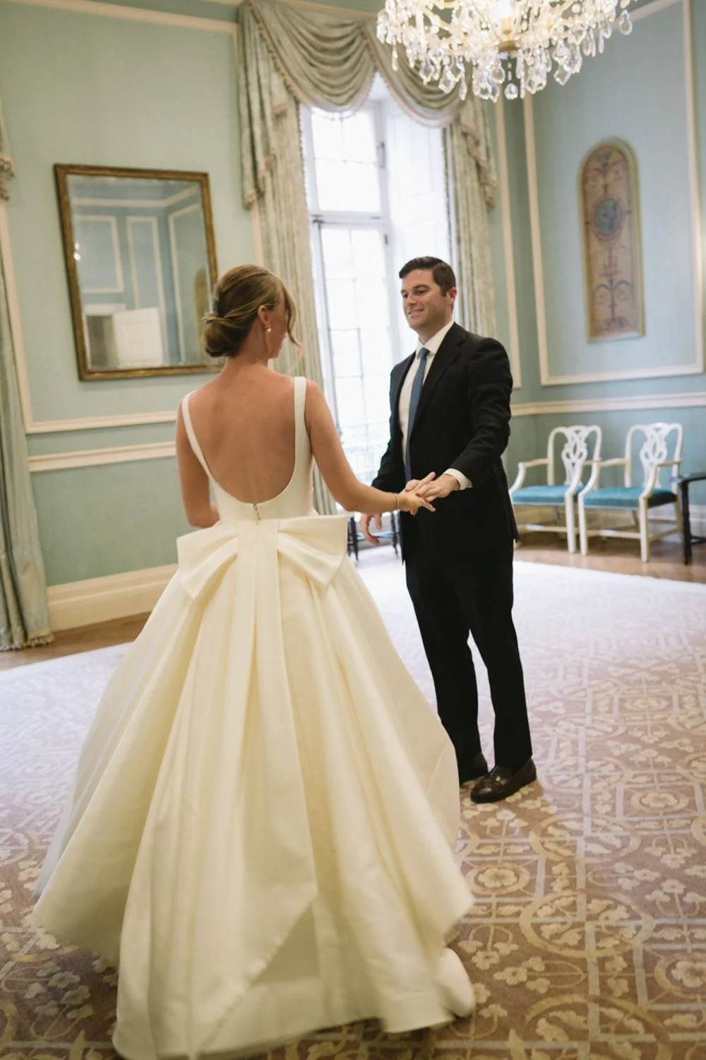A bride and groom holding hands and smiling at each other during a wedding ceremony in an elegant room with green walls, large windows, and a chandelier.