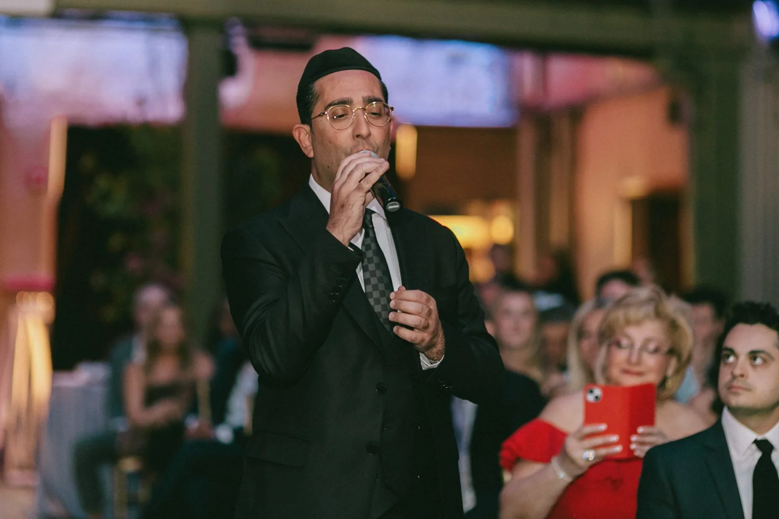 A man dressed in a black suit with a checkered tie and glasses is holding a microphone, speaking in front of a seated audience in an indoor setting.