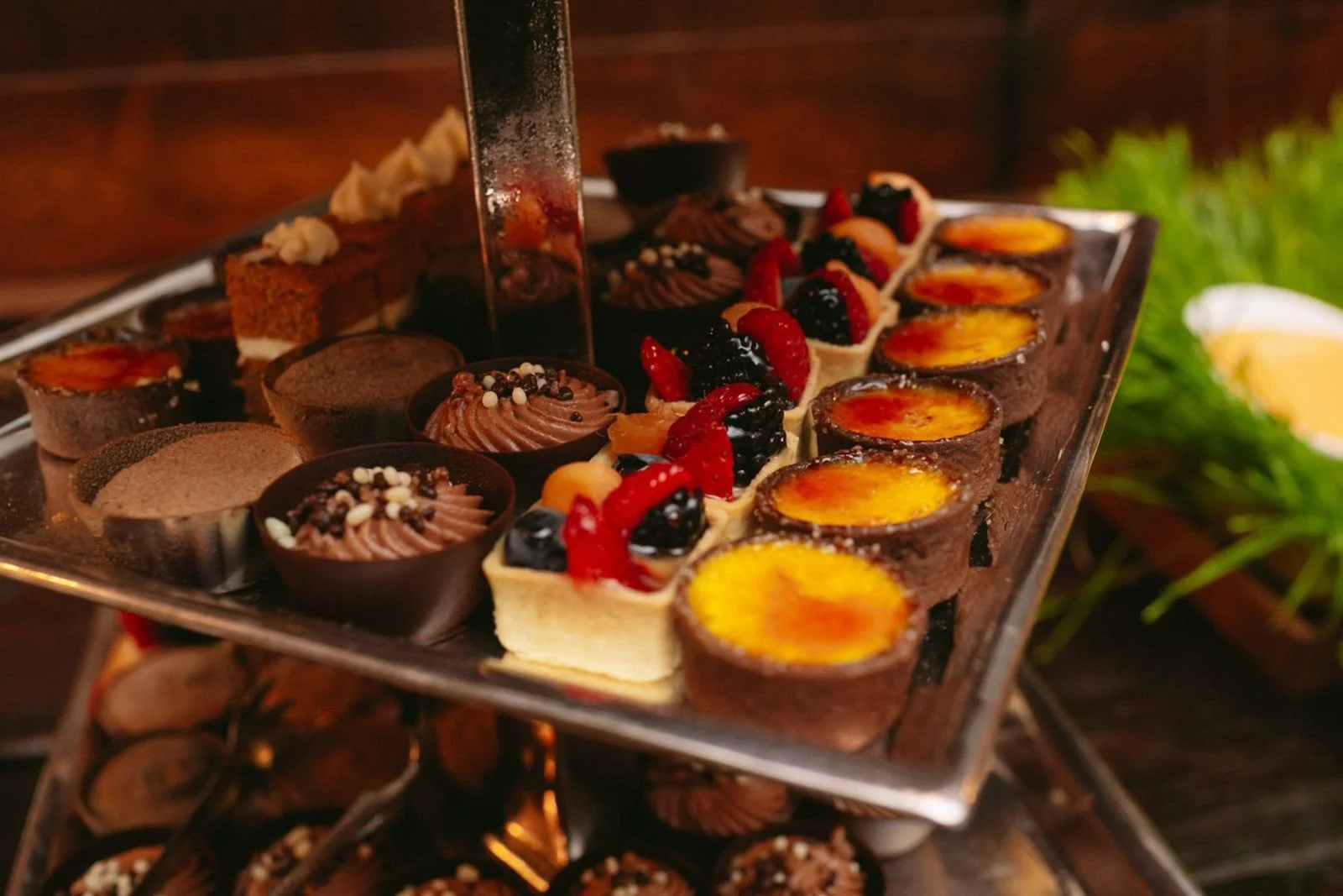 Assorted small desserts on a multi-tiered tray, including chocolate tarts, fruit-topped cheesecakes, and crème brûlée, set against a dark background.