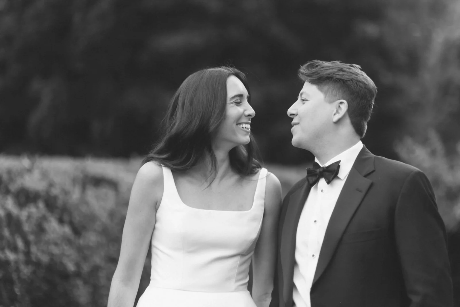 A black and white photo of a woman and a man in wedding attire smiling at each other outdoors.