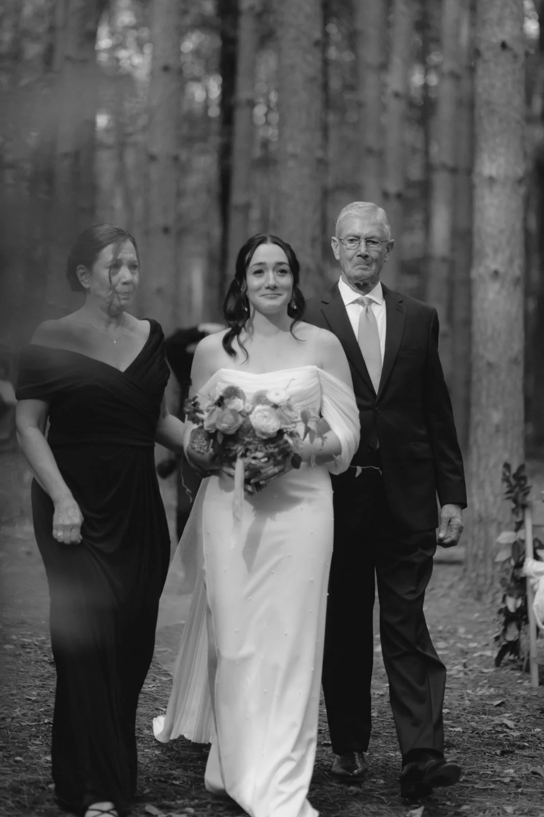 A woman in a wedding dress holding a bouquet, walking down a forest path with an older man in a suit and a woman in a black dress, presumably during a wedding ceremony.