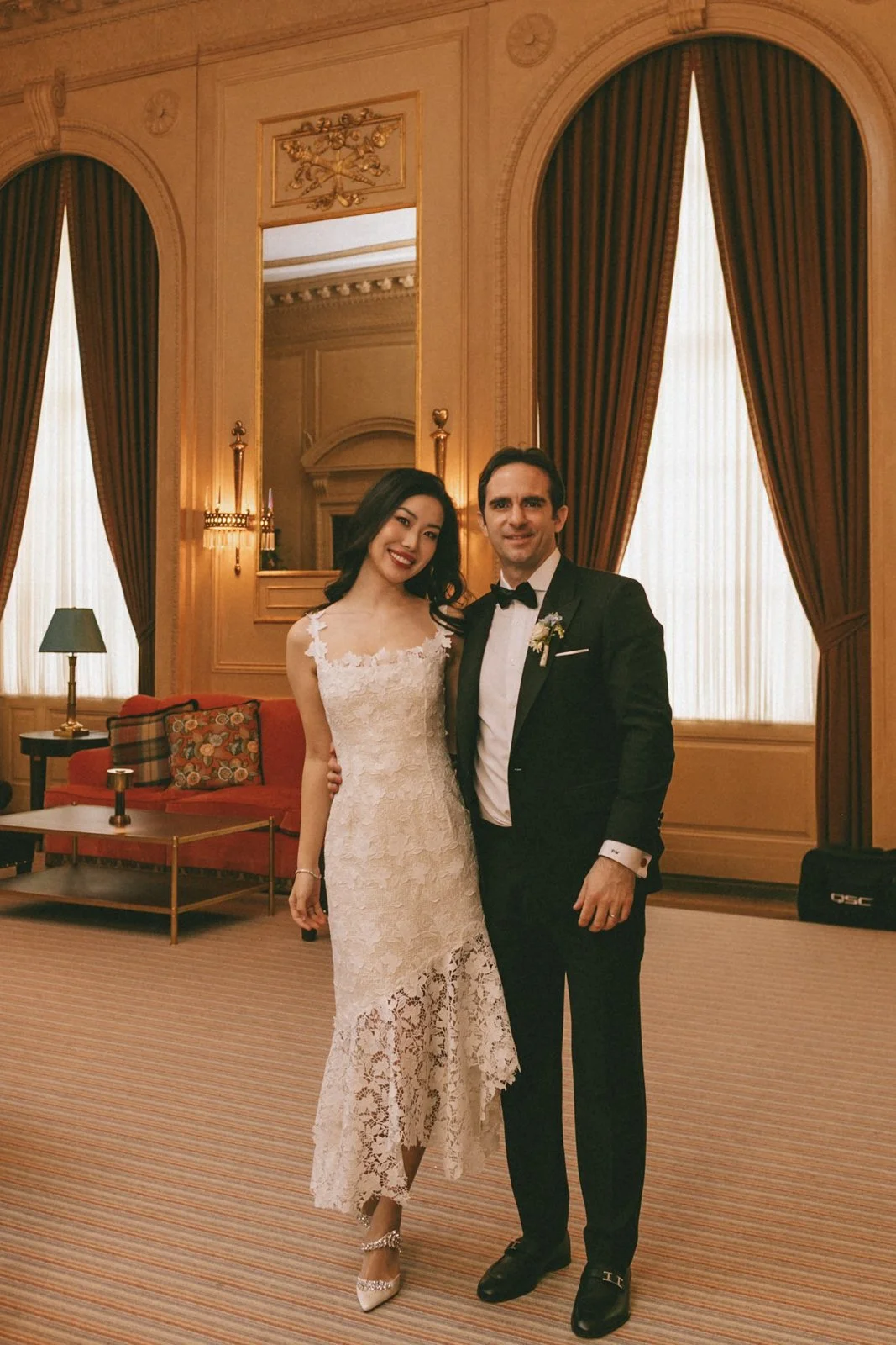 A man and woman dressed in formal attire standing together in an ornate, elegant room with large windows, heavy drapes, and decorative wood paneling.