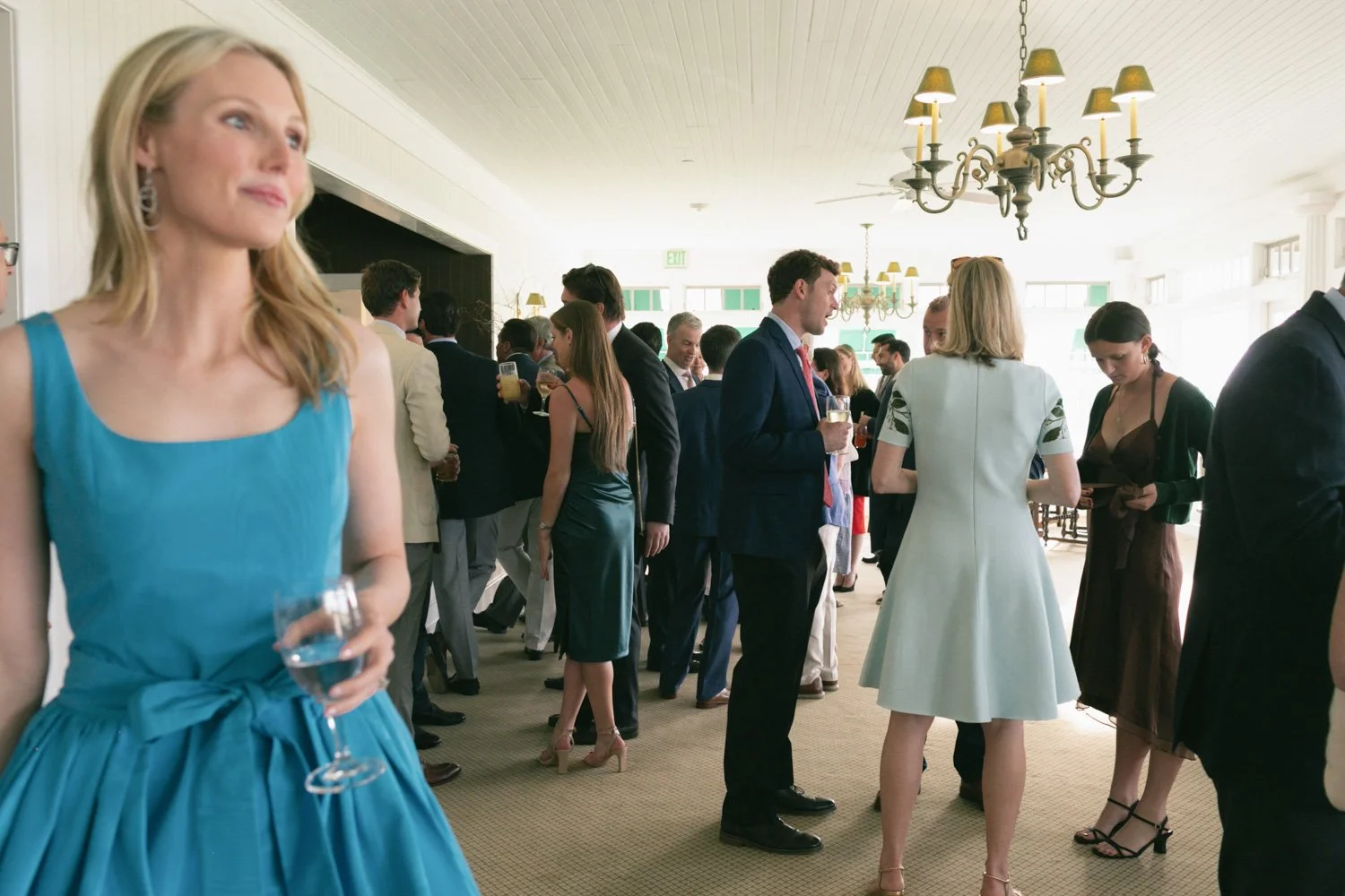 Group of people in formal attire at a social event in a bright, elegant room with chandeliers and large windows.