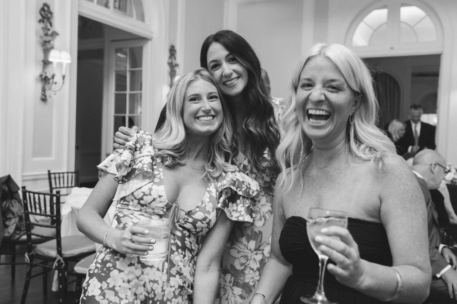 Three women smiling and holding drinks at a social gathering, standing close together in a decorated room with large windows and other guests in the background.