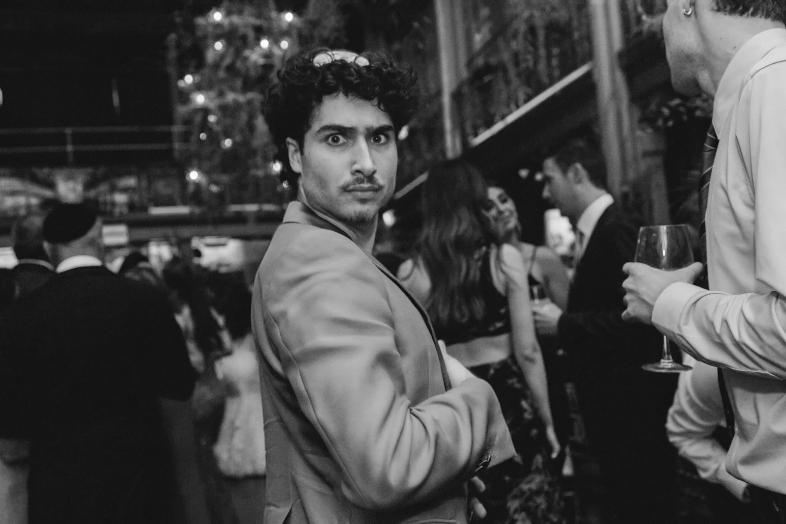 A man with curly hair and a serious expression wearing a suit jacket at a social gathering, surrounded by other people, some holding drinks, in a black and white photo.