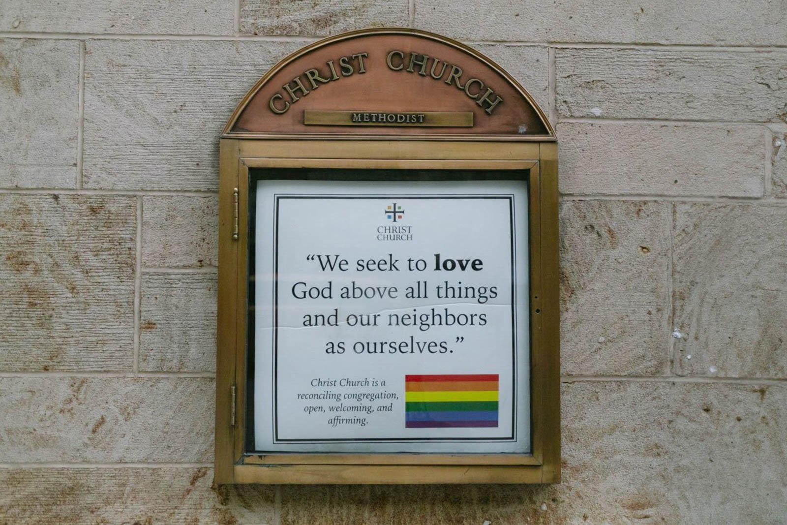 A church display with a quote about love and a rainbow pride flag at Christ Church, labeled Methodist, on a stone wall.