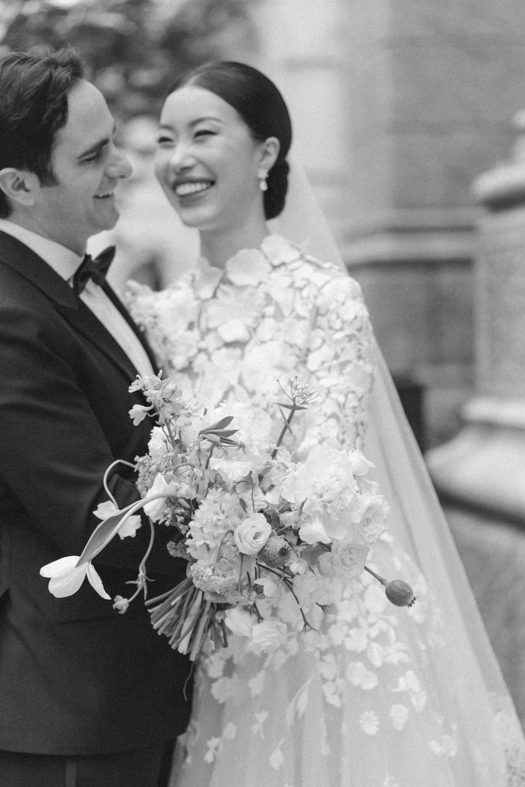A black and white photo of a wedding couple smiling at each other; the groom is in a tuxedo holding a bouquet, and the bride is in a wedding dress with floral details, wearing earrings and a veil.