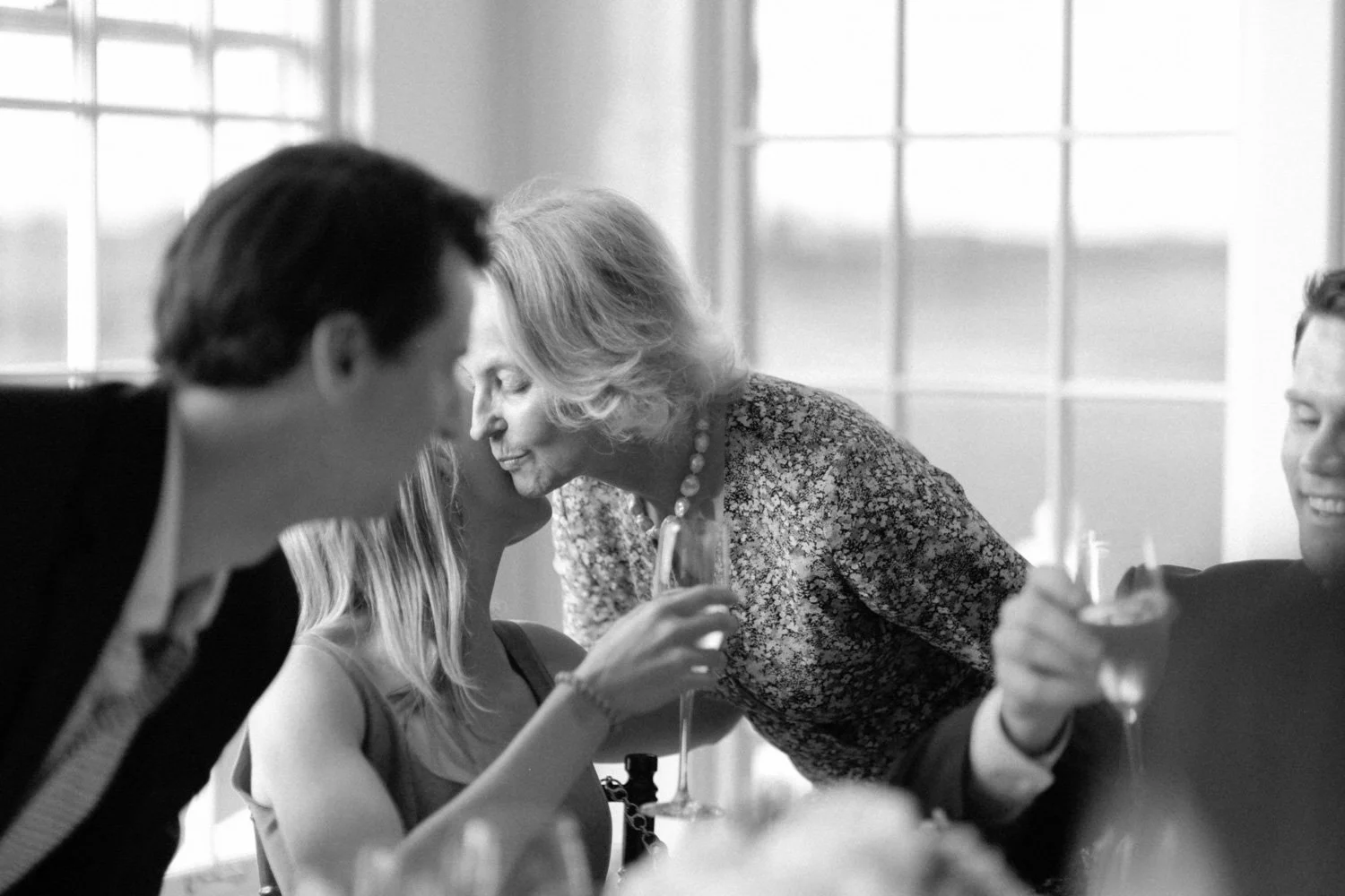 A black and white photo of a social gathering with people engaging in conversation and laughter, with large windows in the background.