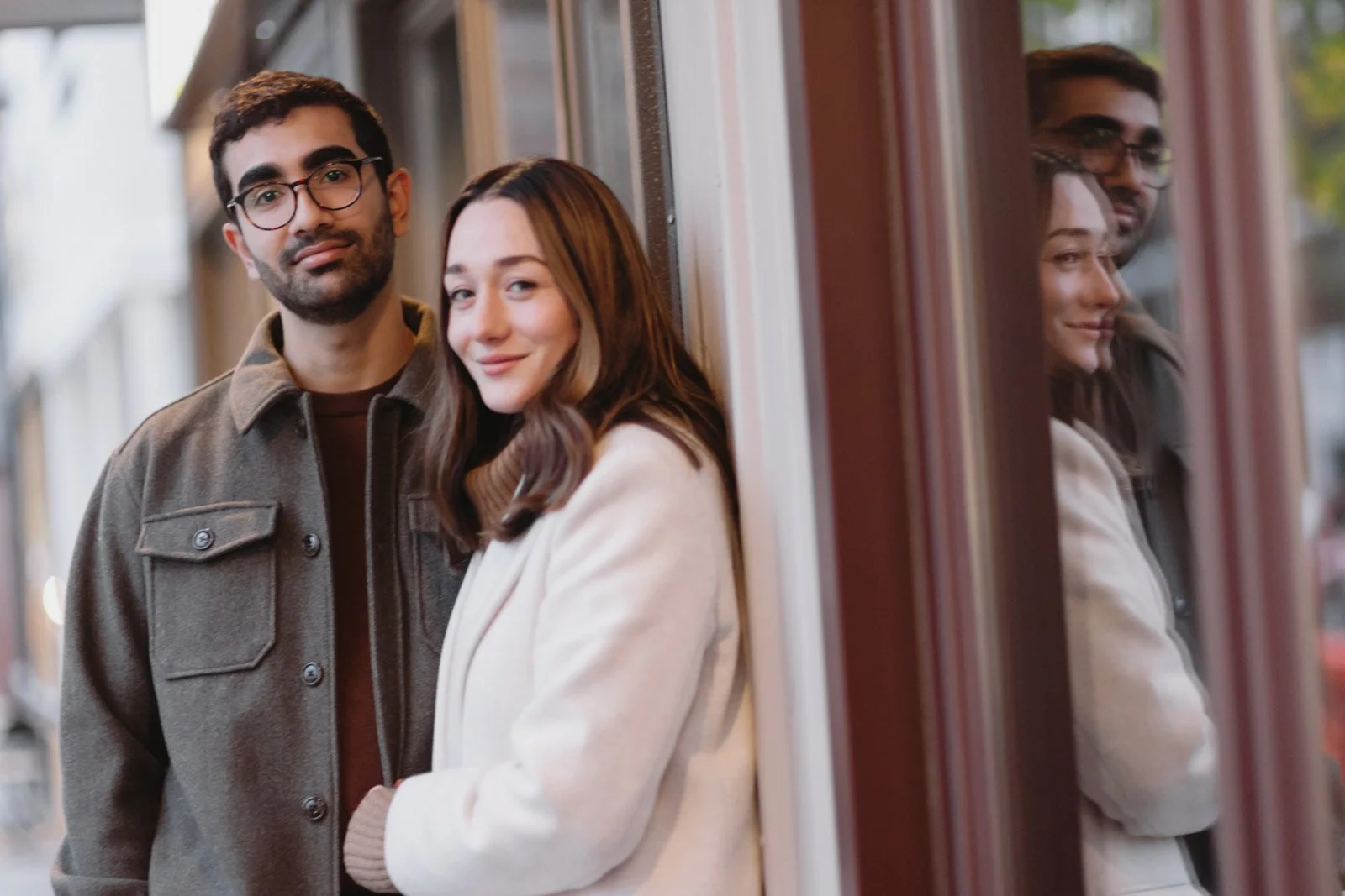 A young man and woman standing close together outside, leaning against a wall with windows. The woman is smiling, and the reflection of both is visible in the window.