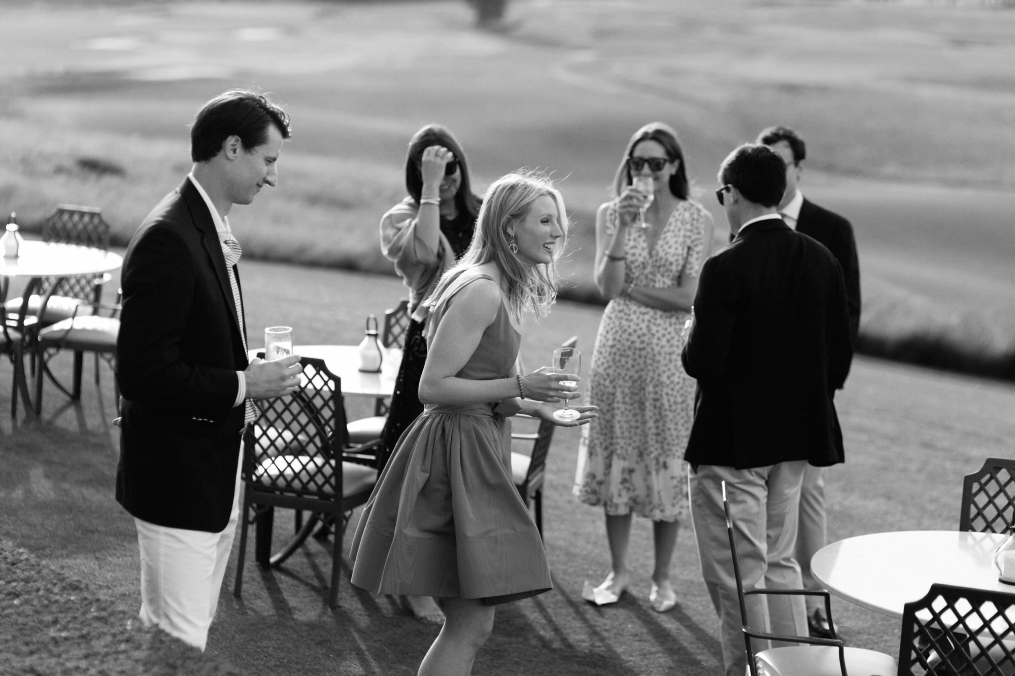 People socializing outdoors, including women and men dressed in semi-formal attire, some holding glasses of drinks, in a grassy area with patio furniture.