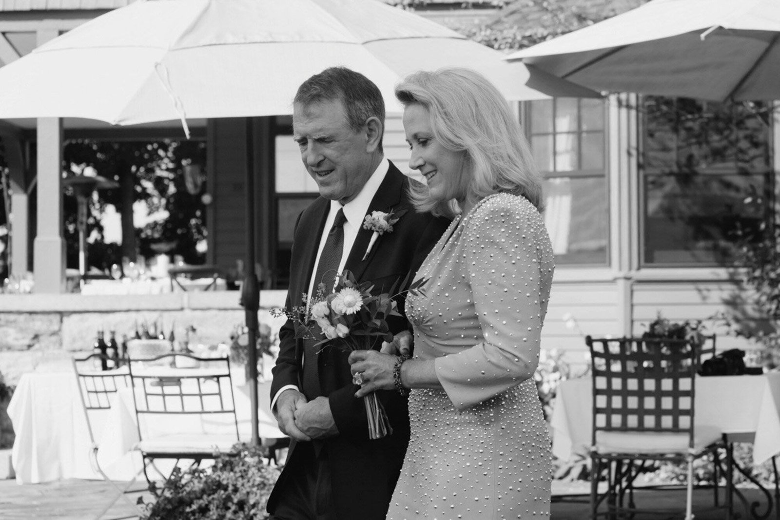 An elderly man and woman walk together outdoors at a gathering, holding a bouquet of flowers, with tables and chairs under umbrellas in the background.